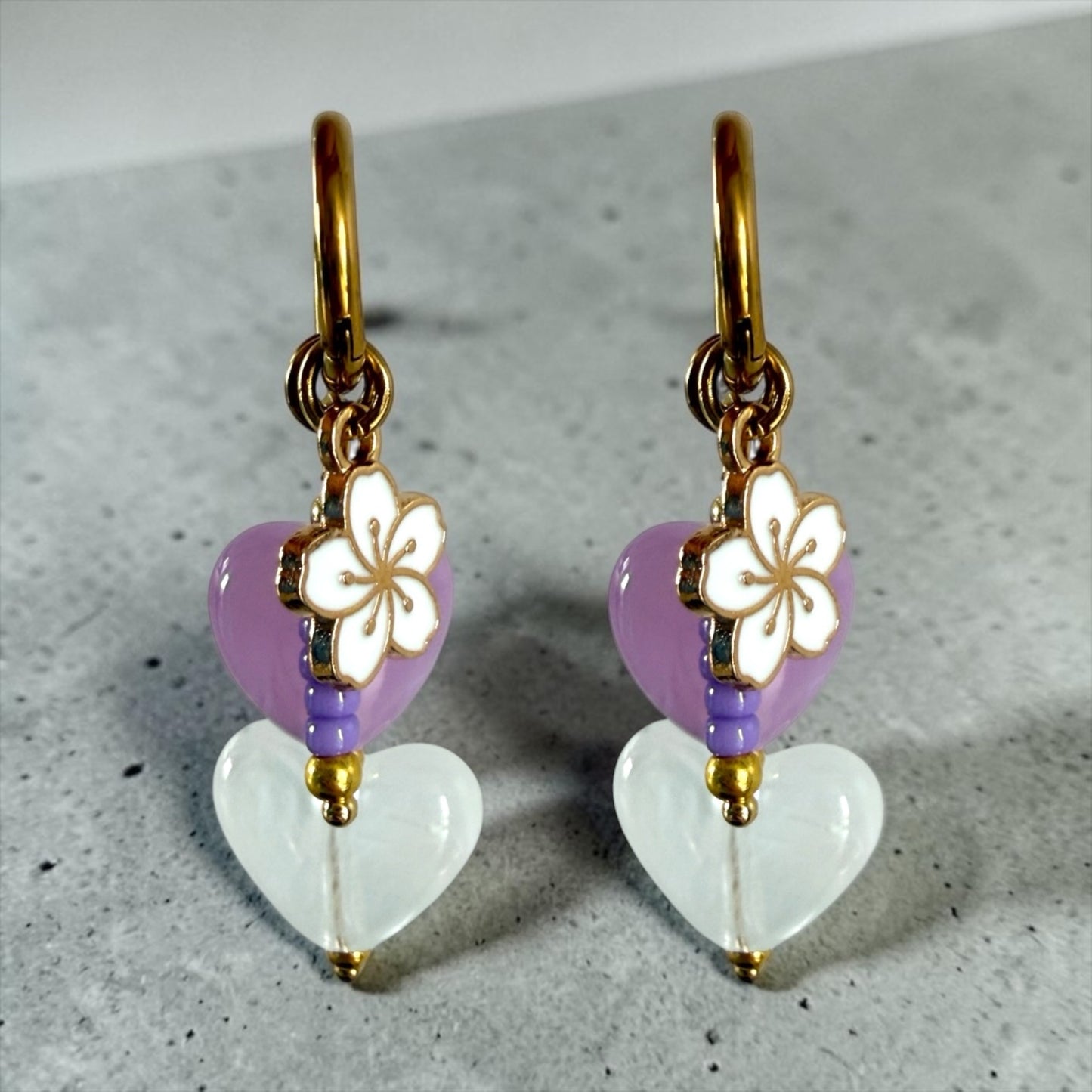 De lavender oorbellen zijn gouden stainless steel oorbellen met een wit en paars hartje, een wit bloemen bedeltje en paarse kralen