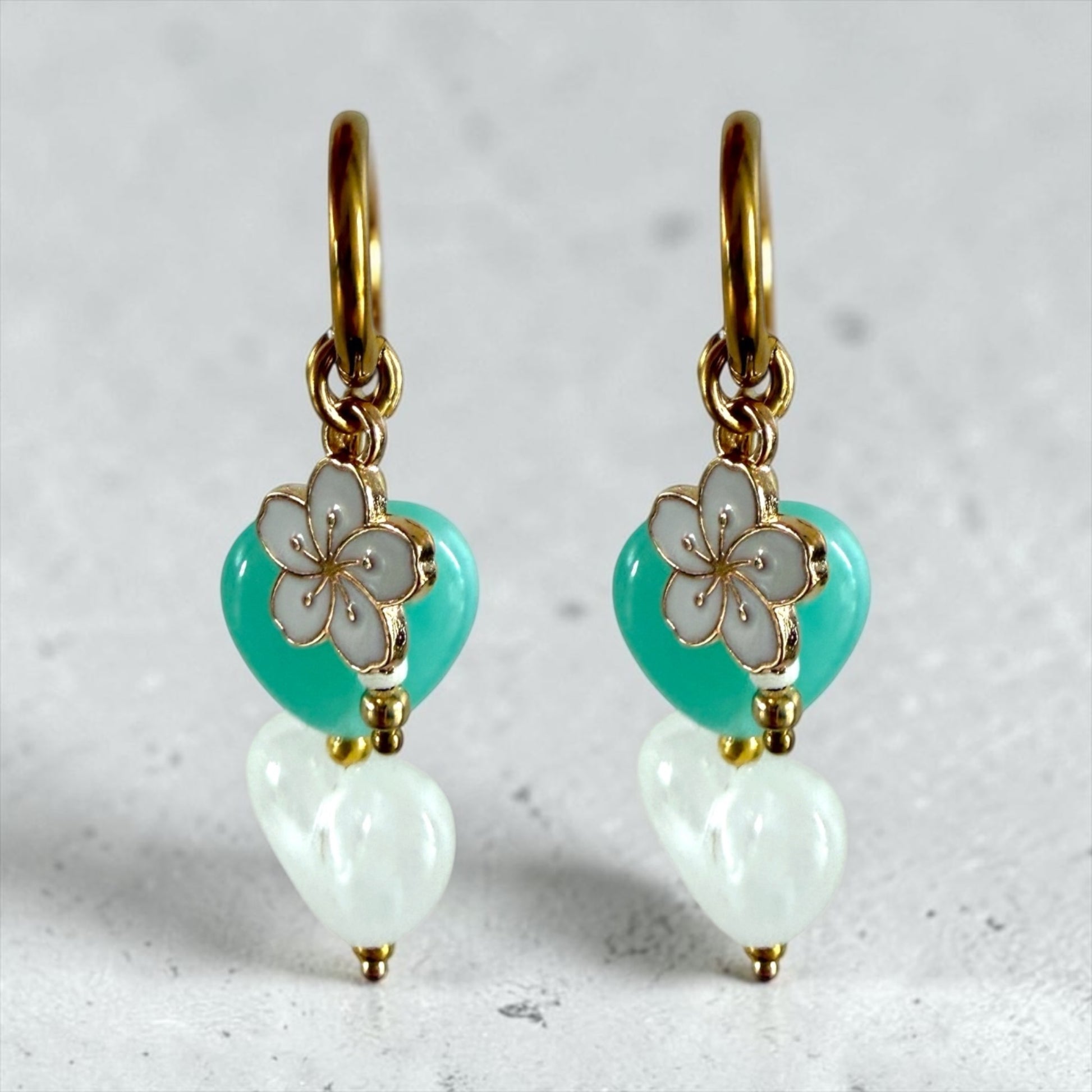 De Lagoon oorbellen zijn gouden stainless steel oorbellen met een wit en turquoise hartje, een wit bloemen bedeltje en witte kralen
