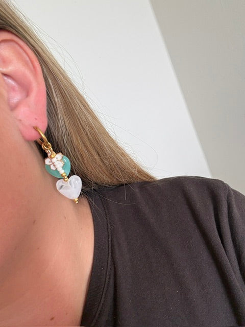 De Lagoon oorbellen zijn gouden stainless steel oorbellen met een wit en turquoise hartje, een wit bloemen bedeltje en witte kralen