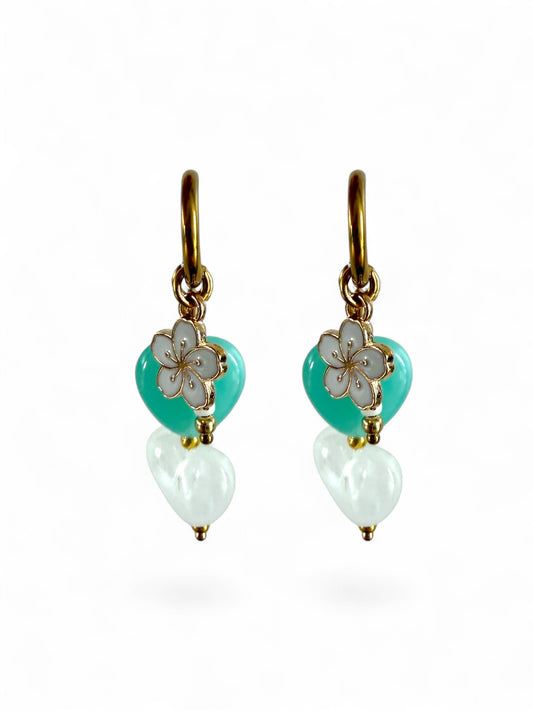 De Lagoon oorbellen zijn gouden stainless steel oorbellen met een wit en turquoise hartje, een wit bloemen bedeltje en witte kralen 
