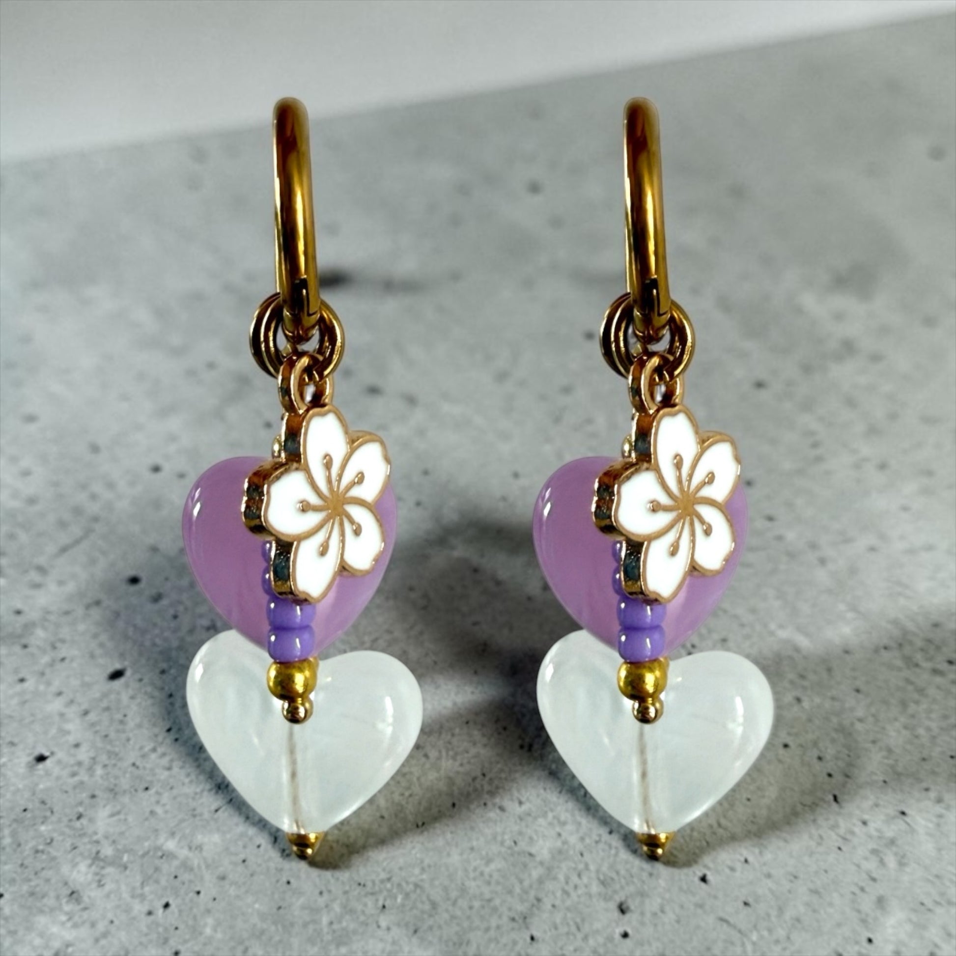 De lavender oorbellen zijn gouden stainless steel oorbellen met een wit en paars hartje, een wit bloemen bedeltje en paarse kralen 