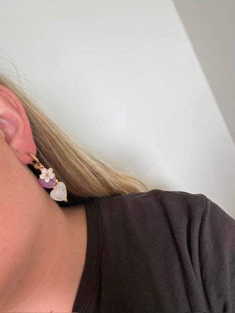 De lavender oorbellen zijn gouden stainless steel oorbellen met een wit en paars hartje, een wit bloemen bedeltje en paarse kralen 